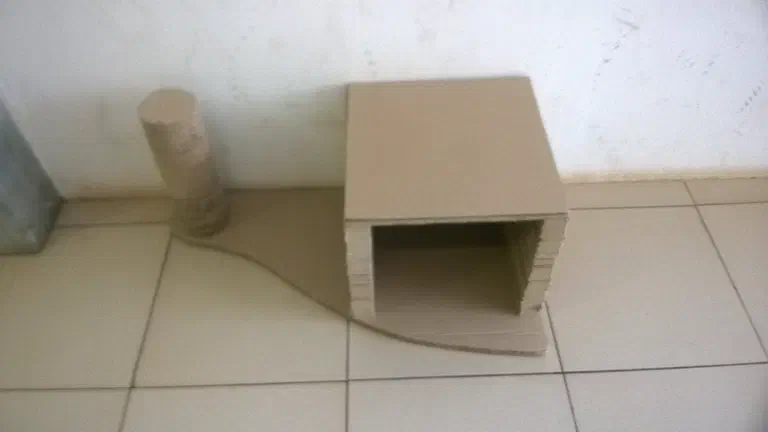Một chiếc nhà mèo tự làm bằng bìa carton đặt sát chân tường, cửa vào khoét hình vòm