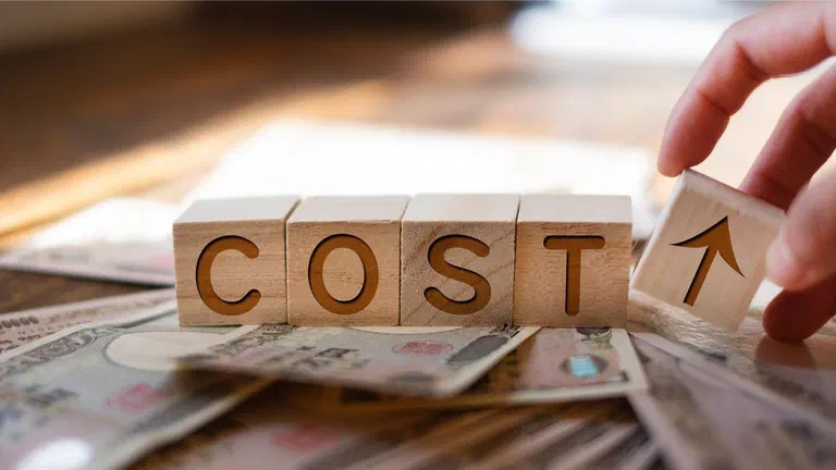 Bàn tay xếp các khối gỗ ghép thành chữ COST kèm khối mũi tên đi lên, đặt trên xấp tiền giấy
